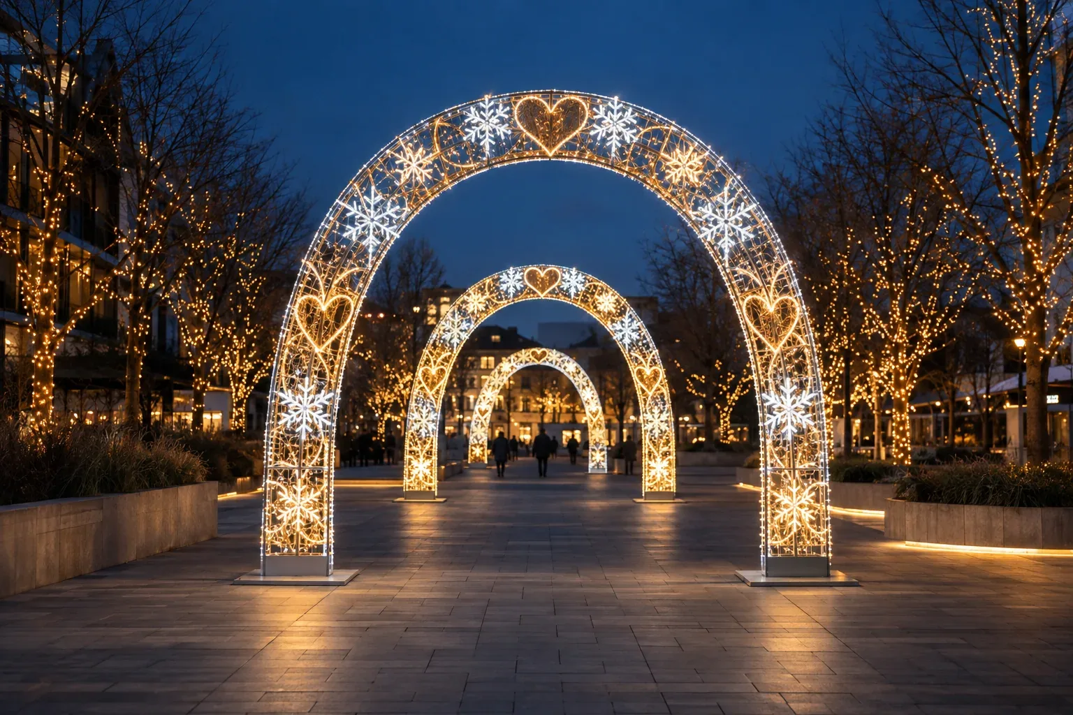 Three lit arches with snowflake and heart shapes along a walkway in a plaza at night