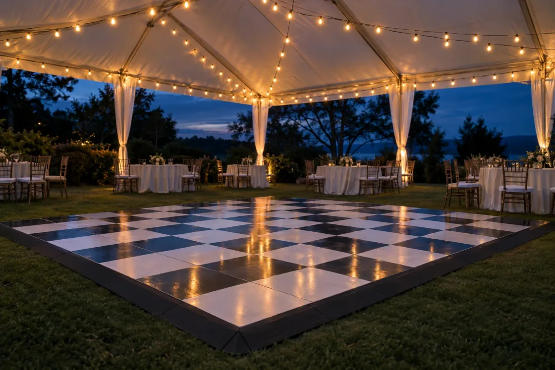 An empty black and white checkerboard dance floor with black edge ramps on the lawn inside a white frame tent at dusk, string lights overhead