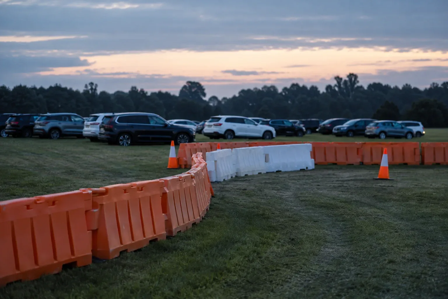 Interlocking orange plastic barricades and a run of white water-filled barriers marking a vehicle lane across a grass parking lot at dusk, with cones and parked cars behind