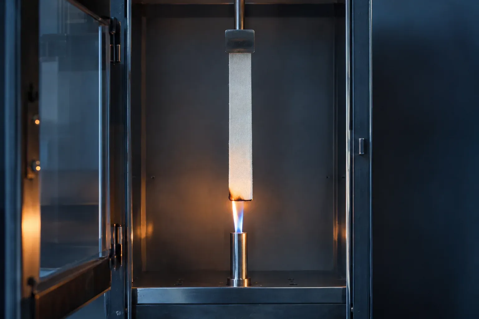 A strip of white coated fabric hanging inside a steel test cabinet, a small blue burner flame at its lower edge
