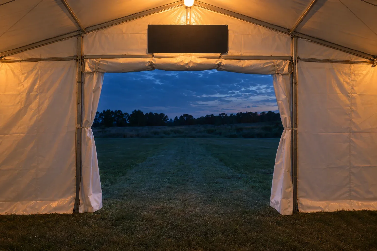 A white tent sidewall rolled up into a wide opening at dusk, a grass path leading straight out and a blank dark sign panel above
