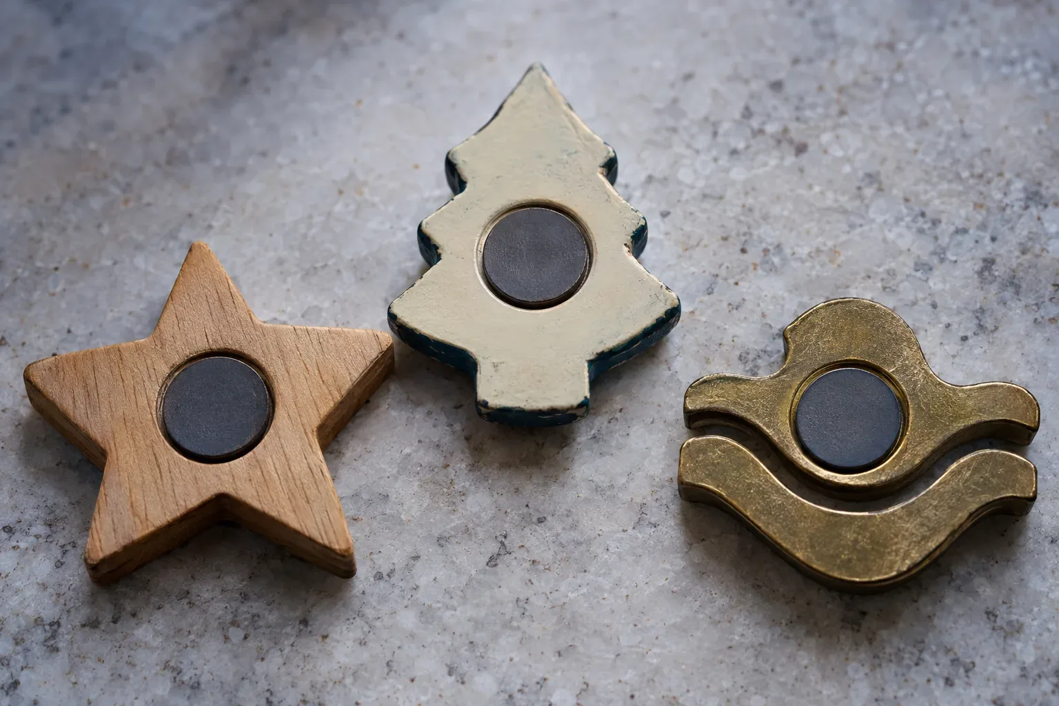 The backs of a wooden star, a cream resin pine tree and a brass wave, each with a round dark magnet seated in a recess