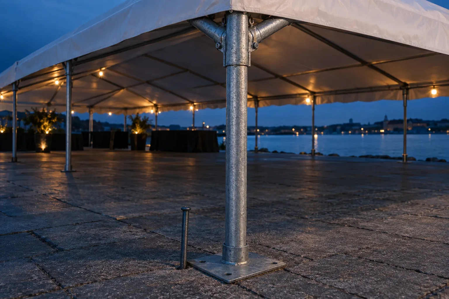 The foot of a galvanized steel tent leg in a square base plate on paving, with the corner fitting joining it to two rafters above