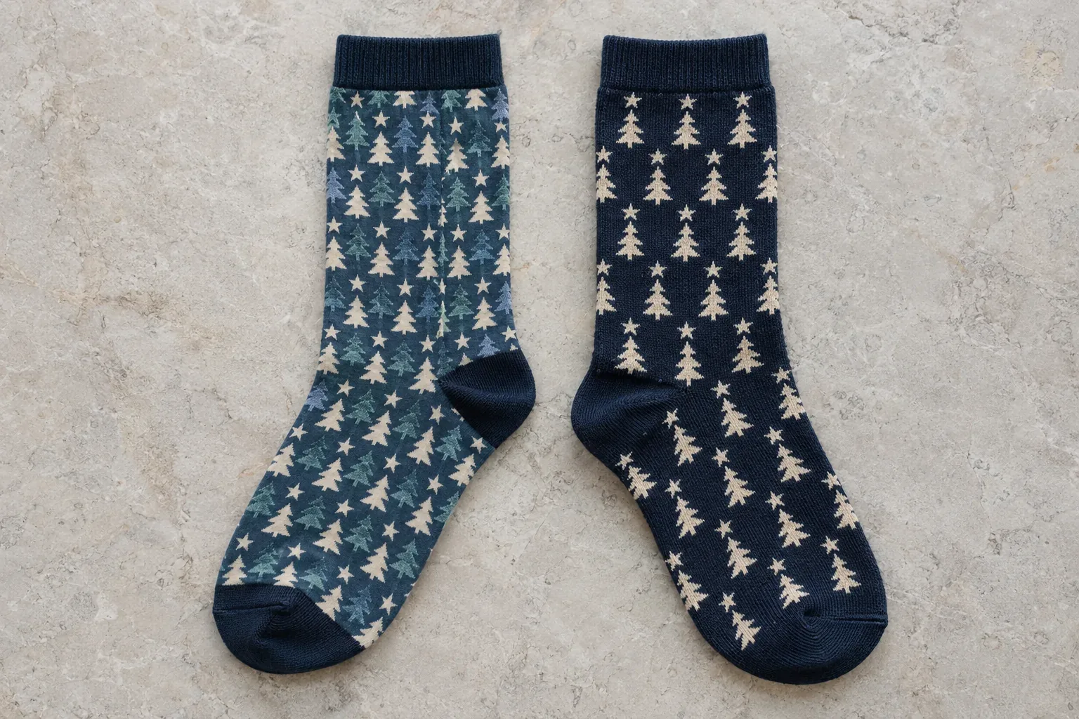 Two socks laid flat on stone: one printed all over with small stars and pine trees in teal and cream, one navy with the same motif knitted in