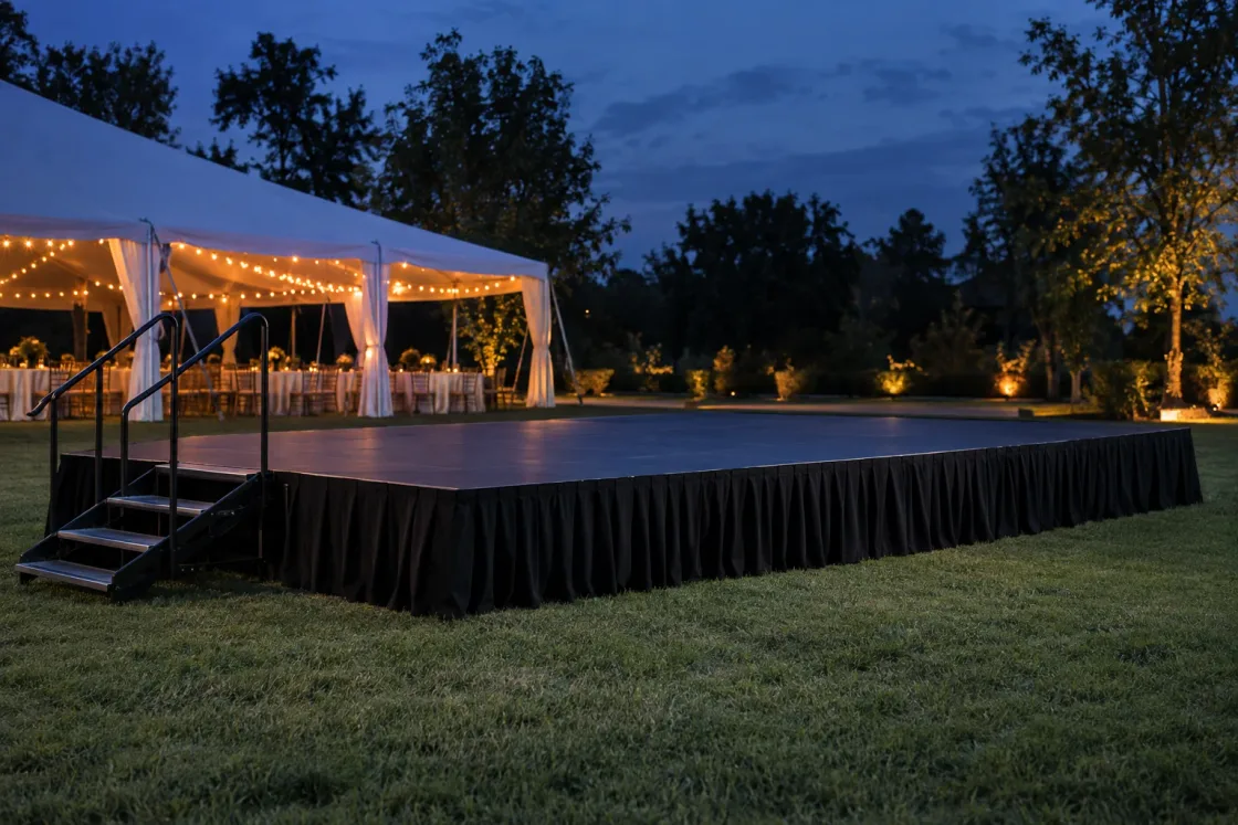 An empty portable stage with a black top and black pleated skirting on a lawn at dusk, steel steps with handrails at one end and a lit white tent behind
