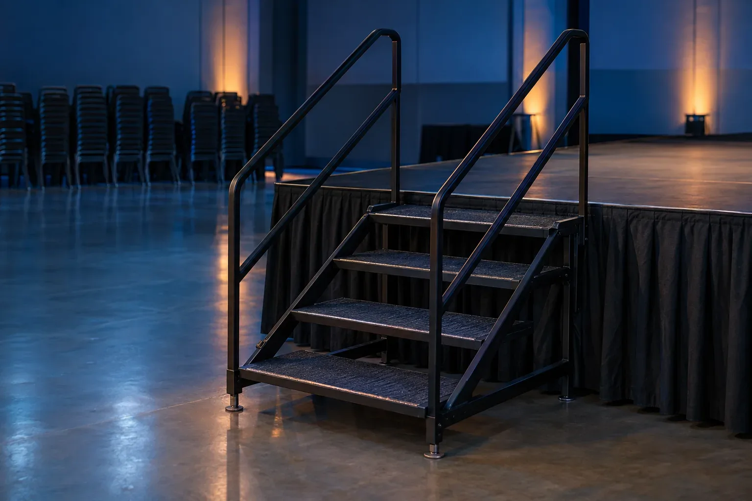 A black steel stair unit with handrails on both sides against the skirted edge of a low stage in an empty hall