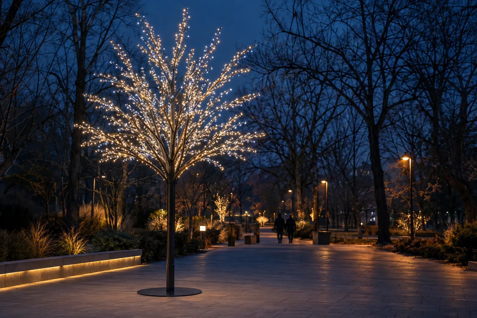 An artificial tree of slender metal branches lined with white lights, on a round base plate beside a park path at night